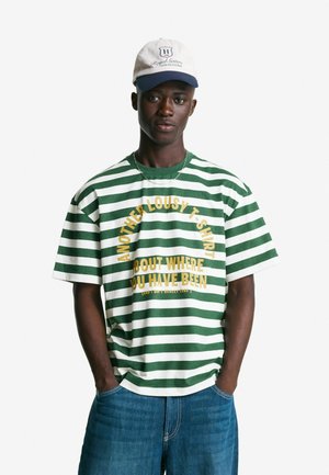 T-shirt a righe verdi e bianche con una vestibilità oversize, con testo dorato che recita "UN'ALTRA INDECENTE T-SHIRT SU DOVE SEI STATO".