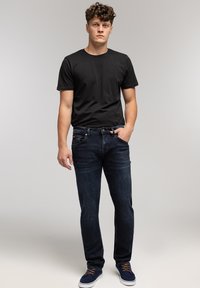 Młody mężczyzna stojący z ręką w kieszeni, ubrany w czarną koszulkę, ciemnoniebieskie dżinsy i granatowe buty sportowe, na jednolitym jasnym tle.