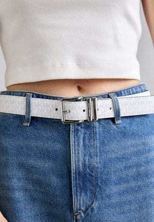Curea albă din piele cu un model embosat, cataramă din metal argintiu, fixată cu bucle pe blugi denim albaștri, asortată cu un top alb cu textura în relief.