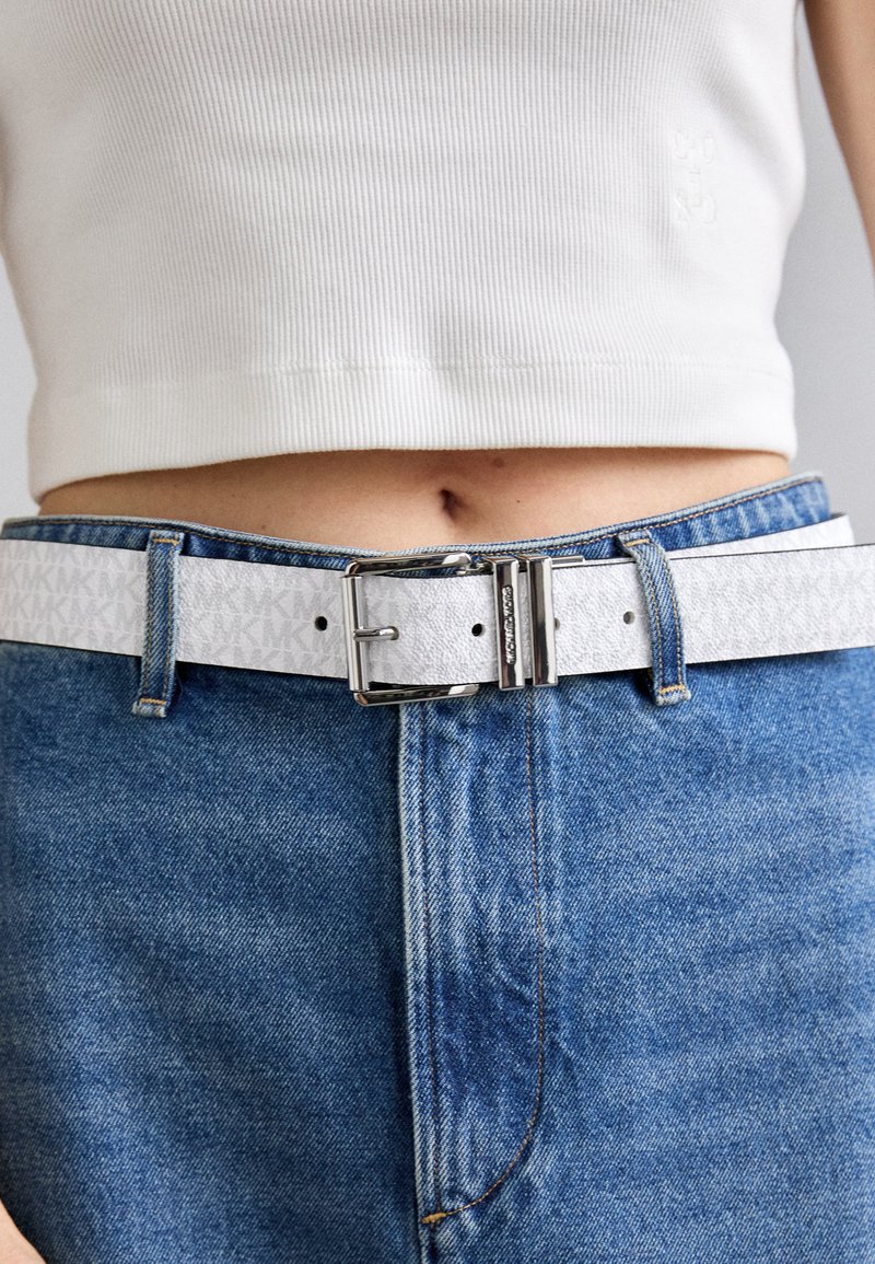 White leather belt with embossed pattern, silver metal buckle, secured with loops on blue denim jeans, paired with a white ribbed top.