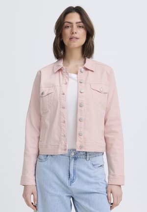 Ung kvinde iført lyserød, knappet jakke over hvid top og lyseblå jeans, stående mod en ensfarvet baggrund.