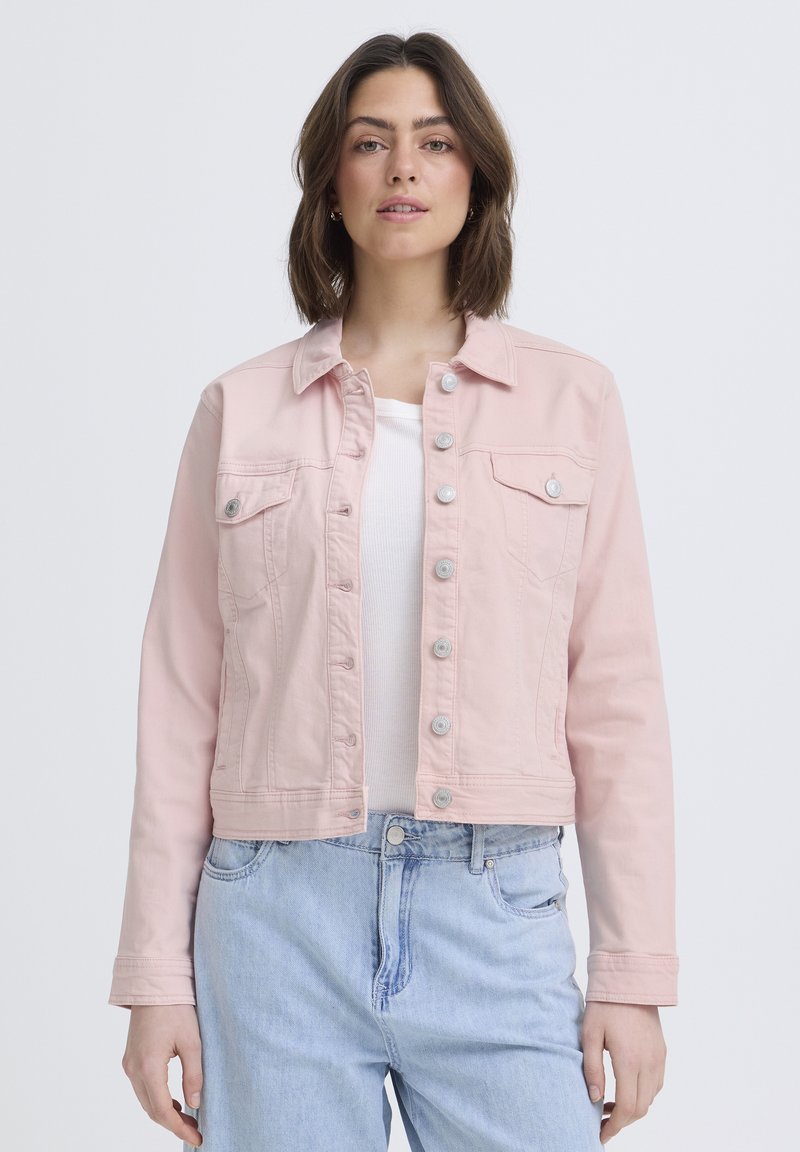 Jauna moteris dėvi šviesiai rožinę užsegamą striukę virš balto marškinėlių ir šviesiai mėlynų džinsų, stovinti priešais vienodą foną.