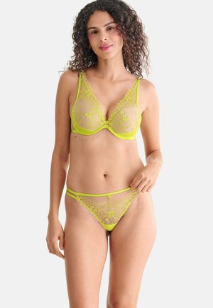 Femme aux cheveux bouclés portant un soutien-gorge en dentelle jaune néon et une culotte en dentelle transparente assortie, debout devant un fond blanc uni.