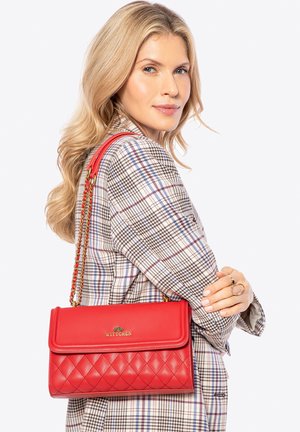 Femme blonde portant un blazer à carreaux tient un sac à bandoulière en cuir matelassé rouge avec une chaîne en or portant l'étiquette "WITTCHEN".