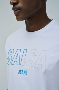 Camiseta de algodón blanca con bordado azul elevado de "SALSA" y "JEANS", cuello redondo, mangas cortas y una textura suave.