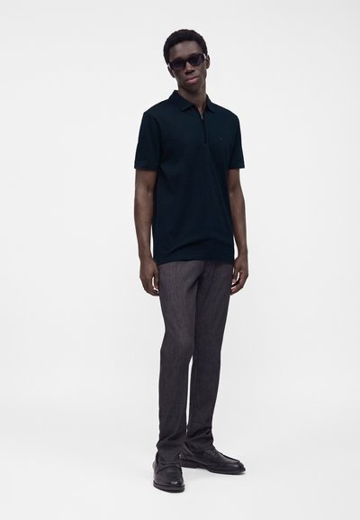 Jeune homme portant des lunettes de soleil noires, un polo noir à manches courtes, un pantalon gris et des chaussures habillées noires, debout devant un fond blanc uni.