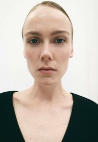 Portrét osoby s hladkou pokožkou, modrými očami a tmavými vlasmi, ktorá má na sebe hlboký čierny odev s výstrihom do V na jednoduchom bielom pozadí.