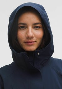 Suurest plaanist naine tumedate juustega, kes kannab lukuga sinist kapuutsiga jope, vaatab kaamerasse neutraalse ilmega.
