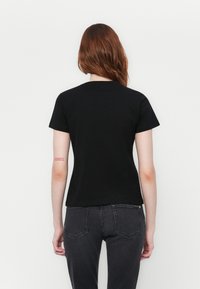 Kvinne med langt rødt hår som vender bort, iført en enkel svart t-skjorte og mørkegrå jeans, stående mot en hvit bakgrunn.