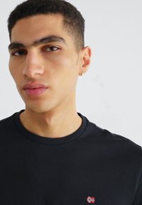 T-shirt en coton noir avec un col rond et des manches courtes, arborant un petit patch brodé rouge, blanc et bleu sur la poitrine.