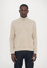 Beige ziphoodie med hög krage och ribbade muddar, med en texturerad tyg och en figurnära design, tillsammans med mörka jeans.