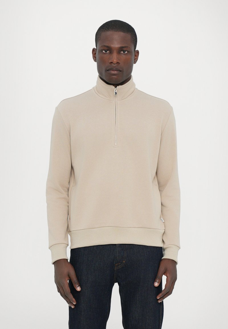 Beige ziphoodie med hög krage och ribbade muddar, med en texturerad tyg och en figurnära design, tillsammans med mörka jeans.