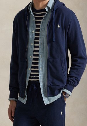 Mand iført navy lynlås hoodie, blå denim knap skjorte, navy og hvidt stribet t-shirt, navy joggingbukser og et armbåndsur.