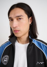 Black zip-up jacket with blue and white accents, featuring a logo on the left chest. Worn over a white T-shirt, textured fabric.