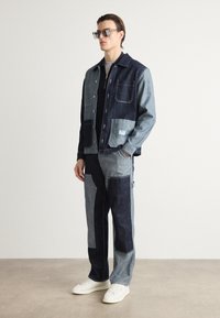 Denim jacket and pants set in varying shades of dark and light blue, featuring patches, pockets, and a relaxed fit. White sneakers accessorized.
