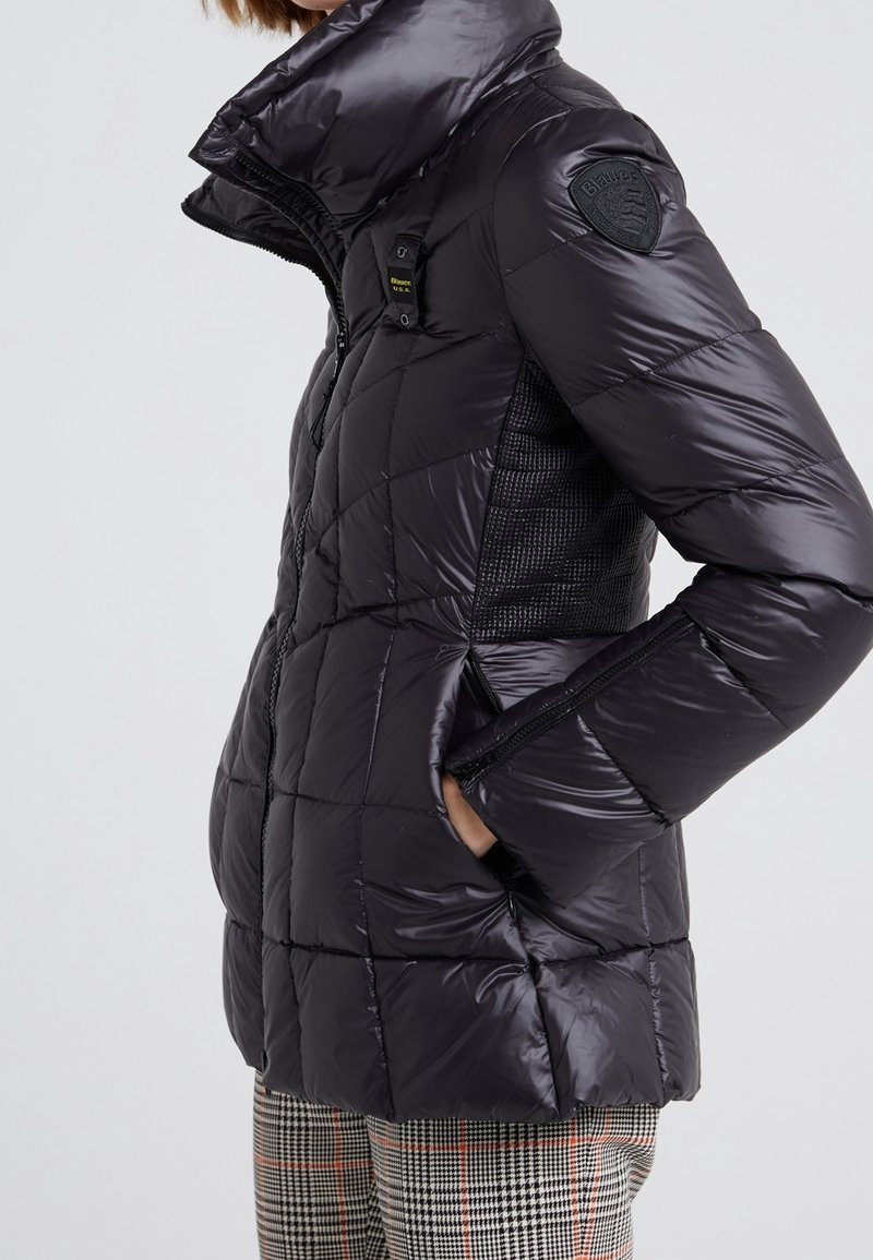 Femme portant une veste matelassée noire brillante avec un col montant, des poches zippées sur les côtés et un écusson embossé sur la partie supérieure de la manche gauche.