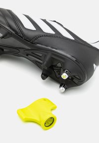 Botín de fútbol negro con rayas blancas y taquetes translúcidos, acompañado de una herramienta amarilla para ajustar los taquetes. Detalle de superficie texturada visible.