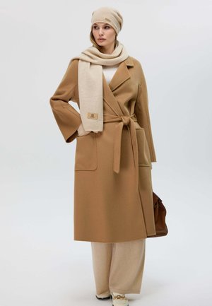 Cappotto avvolgente color tabacco con maniche ampie e cintura, abbinato a una sciarpa beige e un berretto. Pantaloni beige larghi e borsa marrone completano l'outfit.