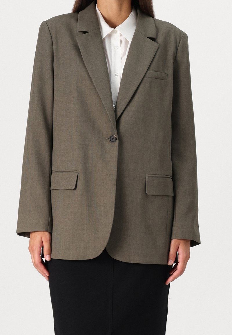 Femme portant un blazer vert olive à un bouton sur une chemise blanche à col avec une jupe noire, debout devant un fond clair uni.