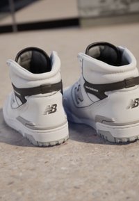 Bílé vysoké tenisky s šedými akcenty, s texturovaným povrchem, vycpaným límcem a výrazným logem "New Balance" na patě.