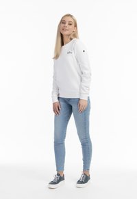Felpa bianca con scollo rotondo e logo cucito sul petto, abbinata a jeans skinny azzurri e sneakers scure.