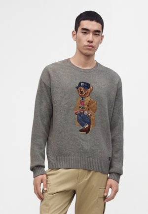 Млад мъж, облечен в сив пуловер с детайлен нашивка на мечка, облечен в спортно облекло и бежови панталони на бял фон.