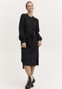 Abito tricot grigio scuro con maniche lunghe e sfiancate, scollo rotondo e vita legata. Presenta delle spaccature laterali ed è abbinato a stivali alla caviglia.