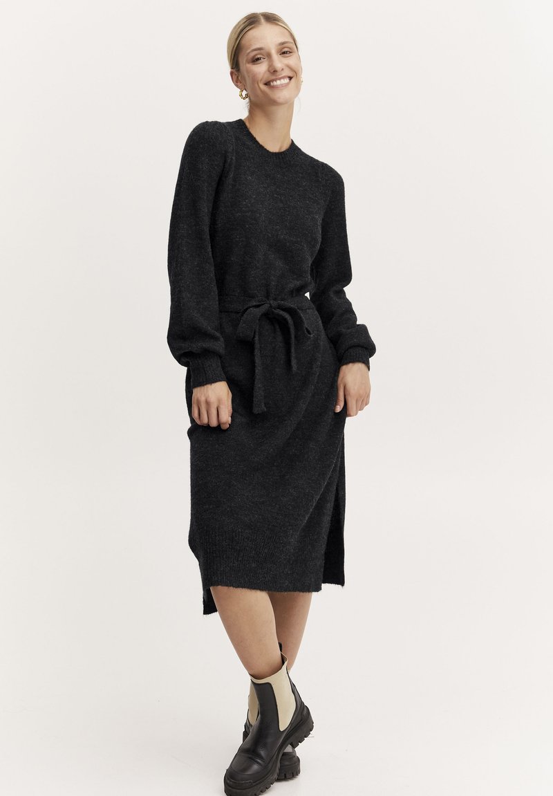 Abito tricot grigio scuro con maniche lunghe e sfiancate, scollo rotondo e vita legata. Presenta delle spaccature laterali ed è abbinato a stivali alla caviglia.