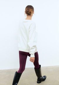 Felpa bianca con polsini e orlo a coste, abbinata a leggings bordeaux e stivali neri fino al ginocchio. Tessuto morbido, vestibilità rilassata.