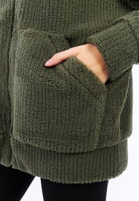 Manteau vert texturé en matériau doux et duveteux ; doté de grandes poches avant et d'un ourlet côtelé, présentant un design douillet.