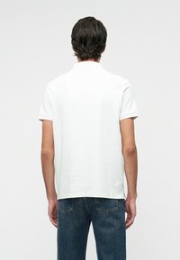 Tricou polo alb cu maneci scurte, cu guler clasic, fabricat din bumbac neted. Previne o croială lejeră și detalii minime de cusătură.