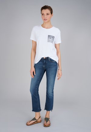 Frau steht, trägt weißes T-Shirt mit Grafik, blaue kurze Jeans mit ausgefransten Säumen und braune Sandalen vor schlichtem grauem Hintergrund.