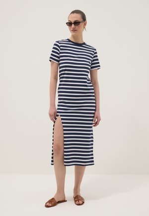 Donna con abito midi a righe blu navy e bianco con spacco laterale, sandali marroni a ciabatta e occhiali da sole neri, in piedi davanti a uno sfondo uniforme.