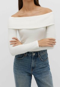 Biały, prążkowany top bez ramiączek z długimi rękawami, w połączeniu z niebieskimi dżinsami z wysokim stanem, posiadającymi zapięcie na guziki i lekkie przetarcia.