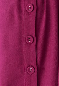 Fuksijos spalvos audinys, ant kurio yra keturi apvalūs mygtukai, kiekvienas su metaliniu kilpu. Tekstūra yra lygi, atrodanti kaip megzta.