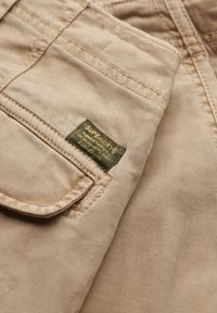 Pantalon en tissu de coton beige avec un gros plan sur une petite étiquette rectangulaire verte cousue près d'un rabat de poche.