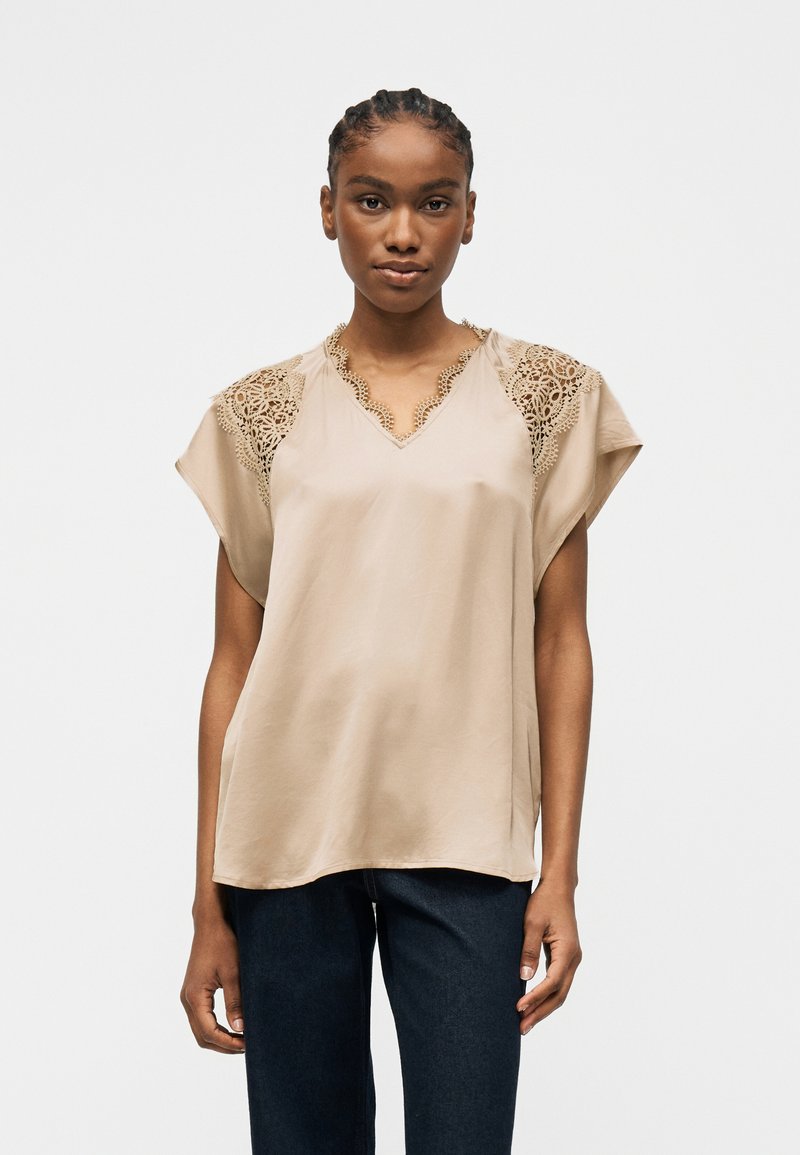 Γυναίκα που φοράει μπεζ μπλούζα με κοντά μανίκια και δαντελένιες λεπτομέρειες στους ώμους και το ντεκολτέ, συνδυασμένη με σκούρα μπλε τζιν, στέκεται μπροστά σε απλό φόντο.