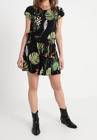 Schwarzes Kleid mit kurzen Ärmeln, gemustert mit grünen Blättern und floralen Akzenten. Elastischer Taillenbund, fließender Rock, kombiniert mit schwarzen Stiefeln.