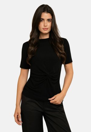 Femme aux longs cheveux foncés portant un haut noir à manches courtes avec un détail froncé sur le devant et un pantalon noir, debout devant un fond uni.