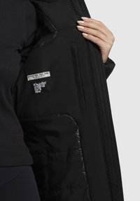Veste isolée noire avec une texture extérieure lisse. Comprend des étiquettes logo et une poche intérieure zippée. Une main tenant la veste montre la doublure intérieure.