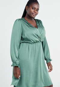 Vihreä kääntömekko, jossa on pitkät, rypytetyt hihat ja joka on valmistettu sileästä kankaasta. Mekossa on v-aukko ja joustava vyötärö, joka luo vartalonmyötäisen siluetin.