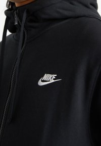 Svart zip-up hoodie i mjukt tyg, med hög krage, framdragkedja och en vit Nike-logotyp på vänster bröst.