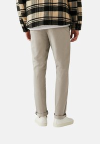 Lichtgrijze chino's, tapered fit, zachte stof, opgerolde zomen, gecombineerd met witte sneakers. De achtergrond is strak wit, zodat de details van de kleding goed uitkomen.