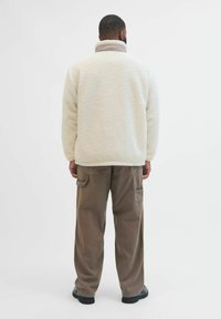 Maglione in pile di colore avorio con colletto alto, abbinato a pantaloni cargo marroni a vestibilità ampia. I materiali testurizzati sono ben visibili.