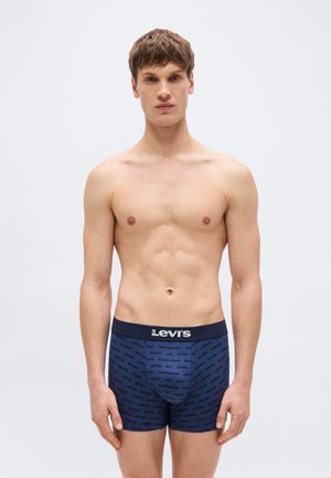 Νεαρός άνδρας που στέκεται με κατεύθυνση προς τα εμπρός, φορώντας σκούρα μπλε μποξεράκια Levi's με μοτίβο λογότυπου σε απλό ανοιχτό φόντο.