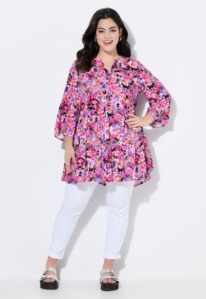 Floral tunic with vibrant pink, purple, and black patterns; features a button front, three-quarter sleeves, and a flared silhouette. Paired with white pants.