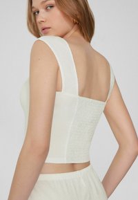Bílý texturovaný crop top s širokými ramínky a čtvercovým designem na zádech, se smockovanými detaily na bocích a hladkým materiálem.