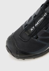 Black athletic shoe with a textured upper, flexible mesh design, and integrated lacing system. Features a reinforced toe cap and branding.