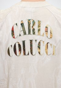 Πίσω μέρος μιας μπεζ μπλούζας με το "CARLO COLUCCI" σε μεγάλα γράμματα γεμάτα με τροπικό φυτό και λουλουδένιο μοτίβο.