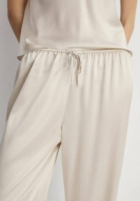 Hlače od svijetle kreme, lagane s elastičnom trakom u struku i detaljem vezanja. Glatka tekstura s bočnim džepovima i opuštenim krojem.